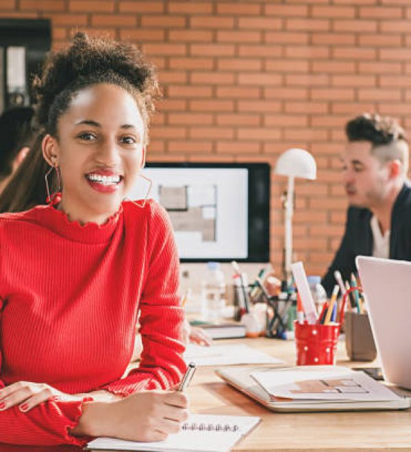 Smiling black business woman in red top holding pen at interior design meeting with office colleagues in casual clothes against brick wall background